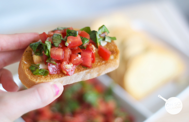 A close up of a hand holding a slice of tomato bruschetta 